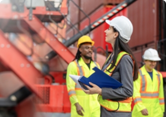 Une superviseure en équipement de sécurité consulte des documents sur un site industriel avec des collègues à l’arrière-plan.